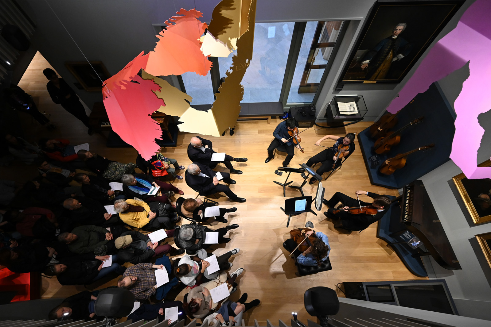 An audience sitting, with a group of musicians playing, looking from above a balcony in a room with historical musical artefacts
